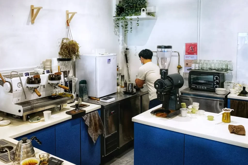 An eye-level, medium shot captures the bustling workspace behind a modern coffee shop counter featuring blue cabinets and stainless steel workstations. On the far left, a professional white espresso machine sits alongside stacked paper cups, a small pitcher, and coffee tamping tools. In the center background, a male barista in a white t-shirt stands with his back to the camera, preparing a drink next to a small white refrigerator. To the right, a large black coffee grinder dominates the foreground counter, which also holds small plastic containers and a gold frothing pitcher, while a toaster oven, glass bottles, and a sink basin line the background under a white wall adorned with hanging plants.