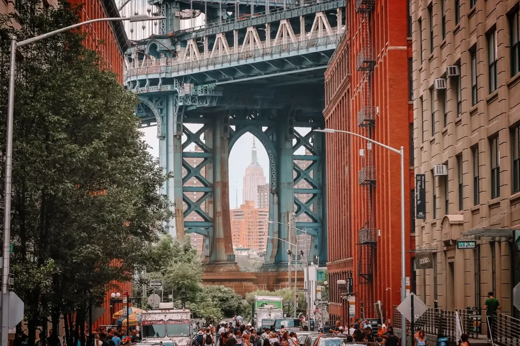 An eye-level, medium-wide shot looks down Washington Street in the DUMBO neighborhood of Brooklyn, capturing the iconic view of the Manhattan Bridge framed by historic industrial buildings. Multi-story red brick structures line the right side of the street, while a mix of brick buildings, green trees, and a street sign reading "FRONT ST" occupy the left. In the background, the massive blue steel tower of the Manhattan Bridge spans across the view, perfectly framing the distant Empire State Building within its lower archway. At street level, a lively crowd of pedestrians and various food trucks fill the roadway, adding a bustling energy to the urban scene.
