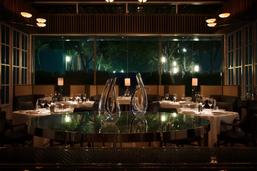 A straight-on, eye-level shot captures a dimly lit, upscale restaurant dining room at night, emphasizing a central, glossy round display table reflecting the room's warm glow. Standing prominently on the mirrored table surface are two large, artistic clear glass wine decanters with sleek, curved shapes. In the background, several dining tables set with crisp white tablecloths, glassware, and small lamps are arranged before a massive, floor-to-ceiling glass window that looks out onto a dark terrace or park with illuminated trees. Elegant wooden paneling and soft, warm overhead light fixtures flank the upper perimeter, creating an intimate, sophisticated atmosphere.
