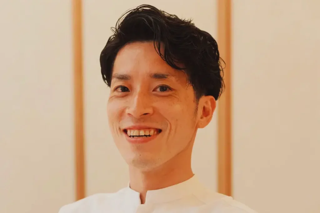 Smiling man with short black hair wearing a white shirt, set against a neutral, softly lit background. His expression conveys friendliness and warmth.