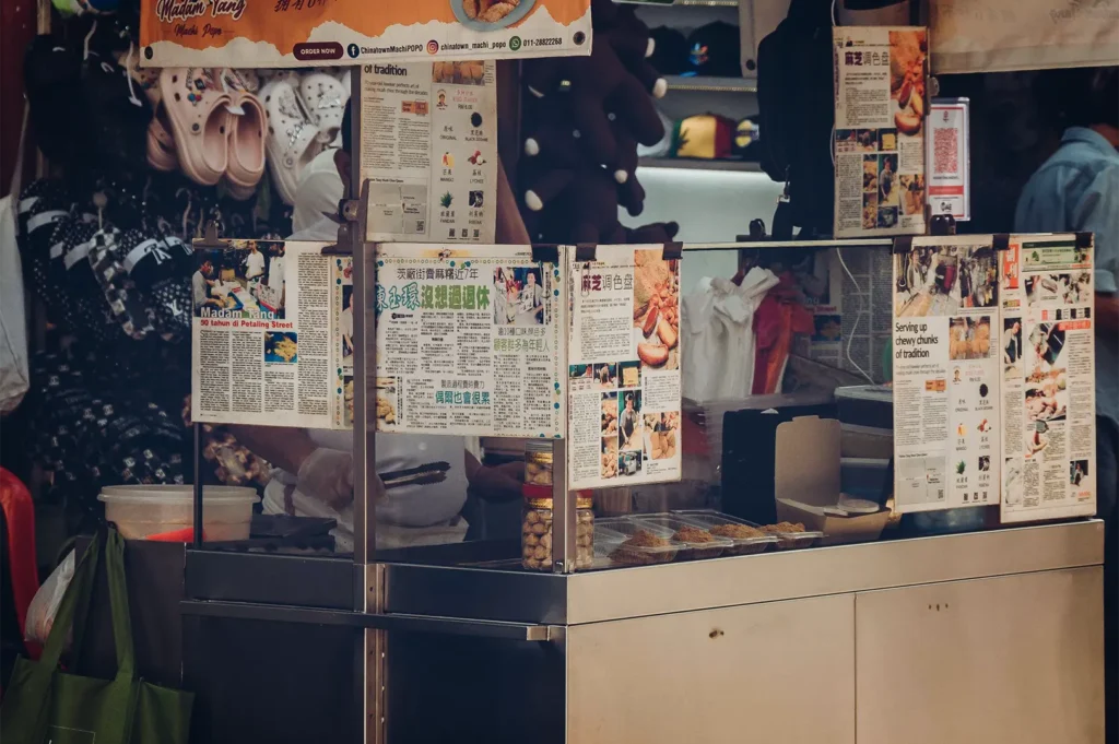 An eye-level shot captures a stainless-steel street food stall decorated with numerous laminated newspaper articles and menus in English and Chinese. Behind the glass panels, a vendor wearing gloves prepares traditional snacks next to clear display trays of ingredients and jars of toppings.