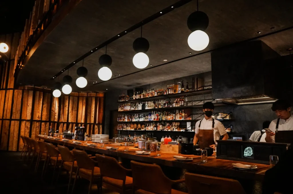 A dimly lit, modern restaurant bar with wooden decor, lined with stools. Round pendant lights hang above. Two chefs prepare behind the bar, evoking a cozy ambiance.