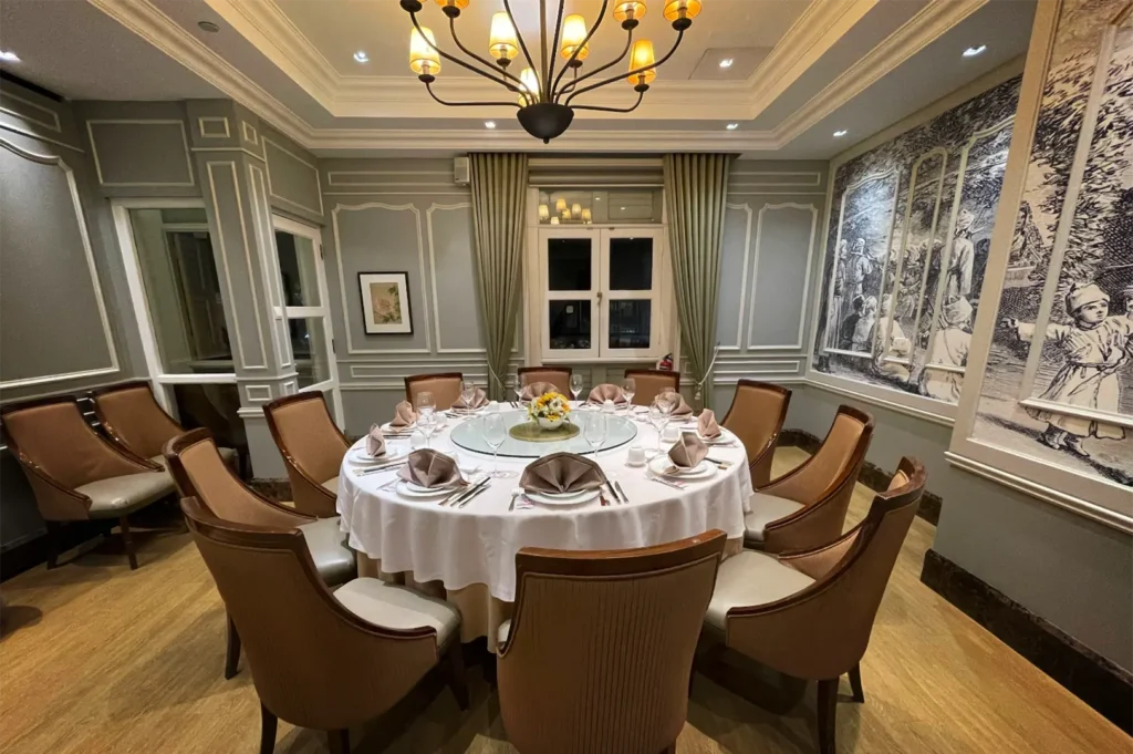 An eye-level shot captures an upscale, private dining room featuring a large round table set with a white tablecloth, formal dinnerware, folded brown napkins, and a lazy Susan centerpiece. The elegant space is detailed with light green paneled walls, a classic chandelier, upholstered high-back chairs, and a large black-and-white illustrated mural depicting historical figures on the right.