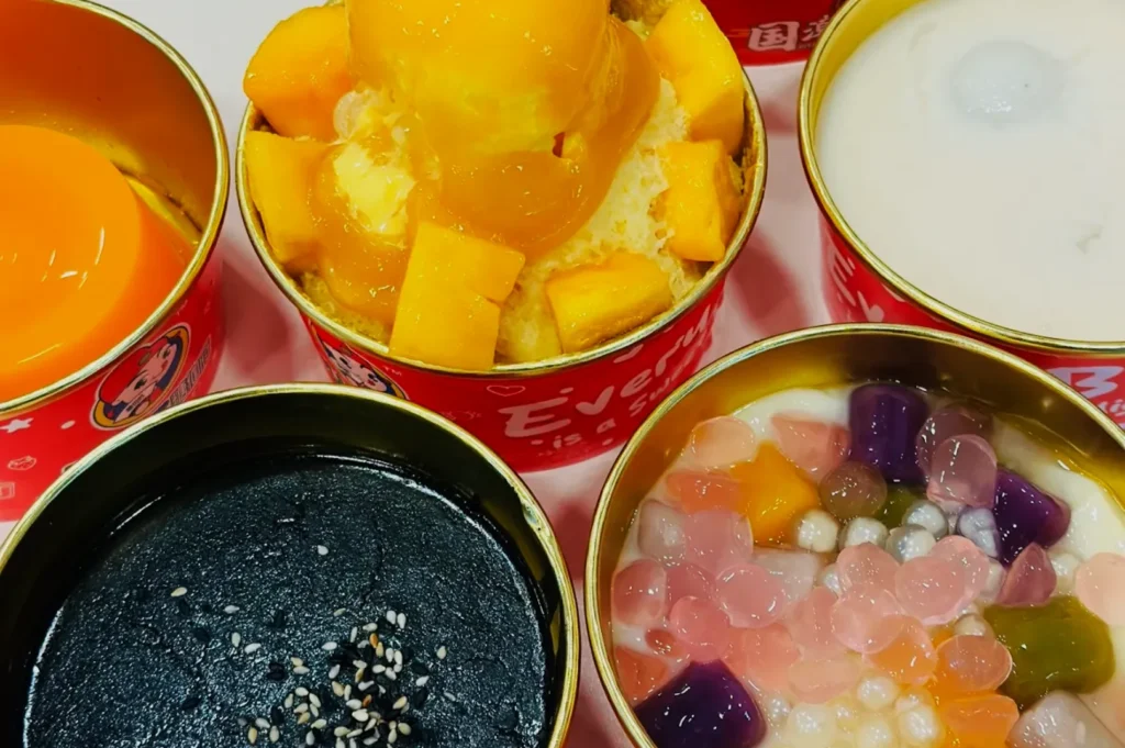 Five colorful Asian desserts in red cups include mango with ice cream, coconut milk, black sesame paste, taro balls, and a pudding.