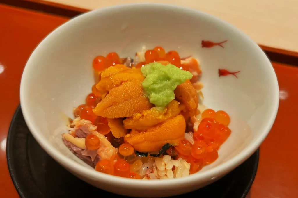 A bowl of rice topped with vibrant orange sea urchin, salmon roe, and a dollop of green wasabi, conveying a fresh and sophisticated culinary experience.