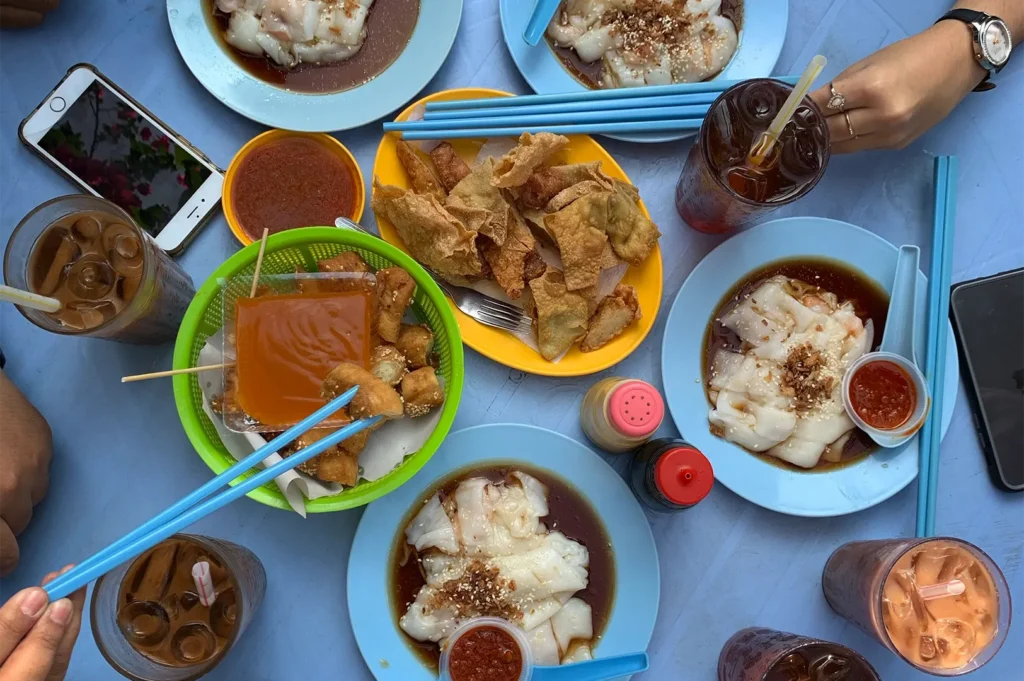 A top-down view captures a light blue table crowded with an assortment of street food, including multiple plates of rice noodle rolls in savory sauce, fried snacks, and several glasses of iced drinks. Around the edges, people's hands are visible interacting with the meal, with one person using bright blue chopsticks to pick up a piece of food.