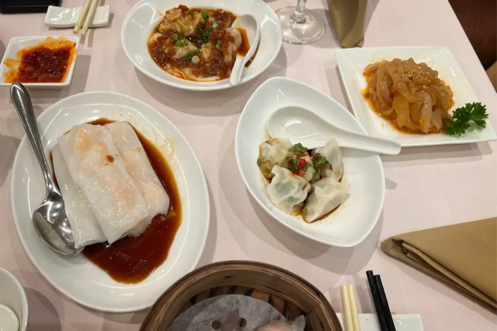 A top-down shot captures a variety of Chinese dim sum dishes arranged on a white tablecloth, including shrimp rice noodle rolls drizzled in sweet soy sauce, savory dumplings, and marinated jellyfish. Spoons, chopsticks, and a small dish of chili sauce flank the plates, alongside the edge of a bamboo steamer basket.