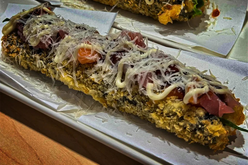 A high-angle, close-up shot captures an elongated sushi roll with a golden, crispy fried exterior served on white parchment paper. The roll is split open and loaded with cubes of raw tuna, drizzles of white sauce, and a generous coating of finely shredded cheese or bonito flakes.