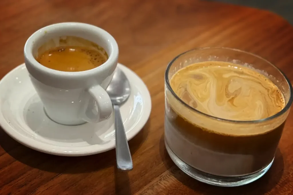 A close-up, slightly high-angle shot captures two different coffee drinks served side by side on a smooth, warm-toned wooden table. On the left, a small, thick white ceramic espresso cup filled with a dark espresso showing a rich, golden-brown crema sits on a matching white saucer with a small silver spoon resting beside it. To the right, a clear, handleless glass displays a layered coffee beverage, featuring a visible gradient from a white milk or cream base at the bottom to a deeper brown espresso layer, topped with swirling patterns of light brown crema and milk across the surface.