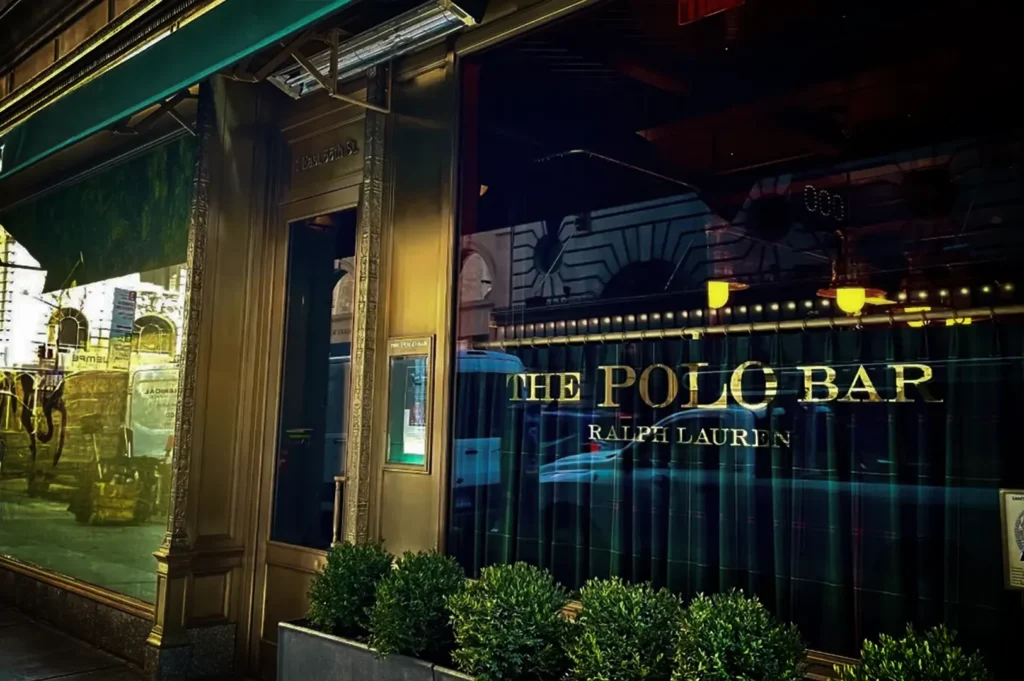 An eye-level, medium shot captures the sophisticated exterior facade of "THE POLO BAR" by Ralph Lauren on a city street. The restaurant entrance features polished brass framing, a dark glass window displaying the establishment's name in bold gold lettering, and a deep green tartan plaid curtain lining the lower half of the window. A row of neatly manicured green shrubs sits in a planter box along the sidewalk in the foreground, while a green awning with "RALPH LAUREN" text is visible on the far left, and a blue "Grade A" sanitary inspection sign stands on the far right.