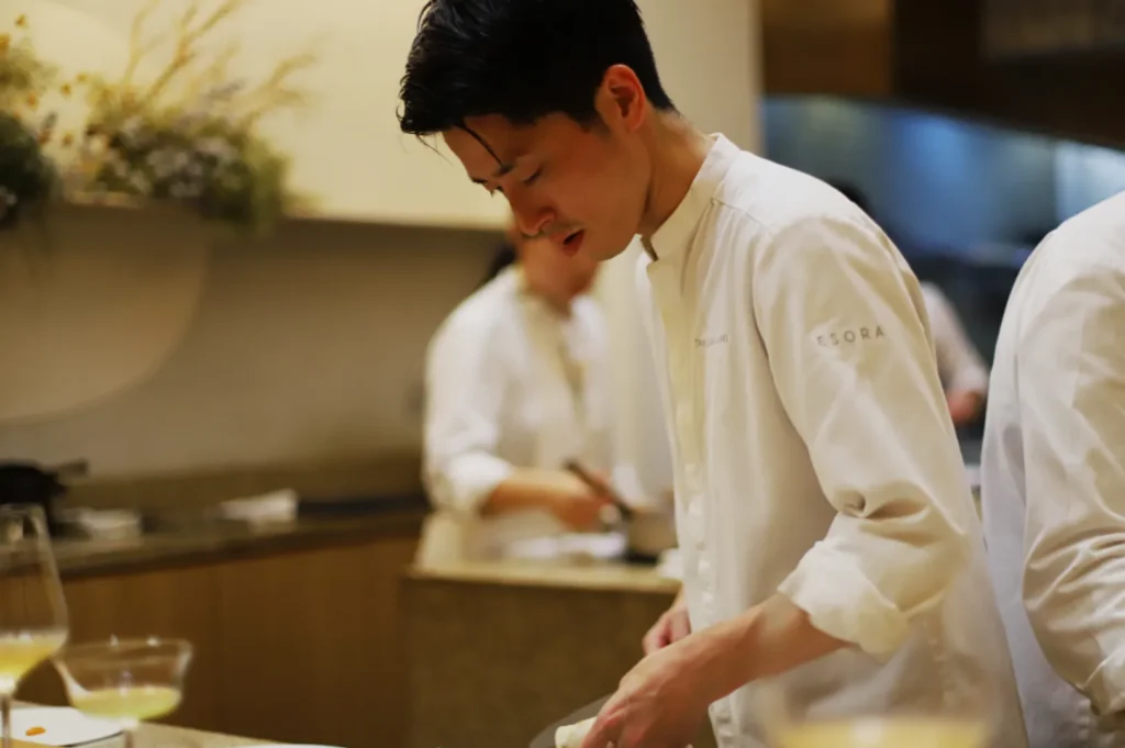 Chef focused on cooking in a professional kitchen. Wearing a white chef's jacket, he works with precision and concentration. Warm lighting creates an inviting atmosphere.