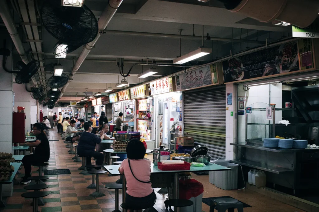 An eye-level shot captures the narrow walkway of an indoor hawker center lined with traditional food stalls featuring illuminated Chinese and English signage. Patrons sit on fixed round stools at small tables beneath industrial ceiling fans and exposed pipes, creating a bustling, dim dining atmosphere.
