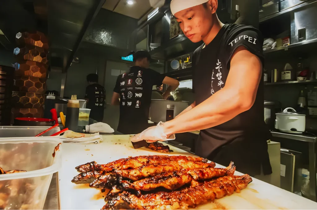 A chef wearing a black shirt and white headband prepares grilled eel on a cutting board in a bustling kitchen, conveying focus and culinary artistry.