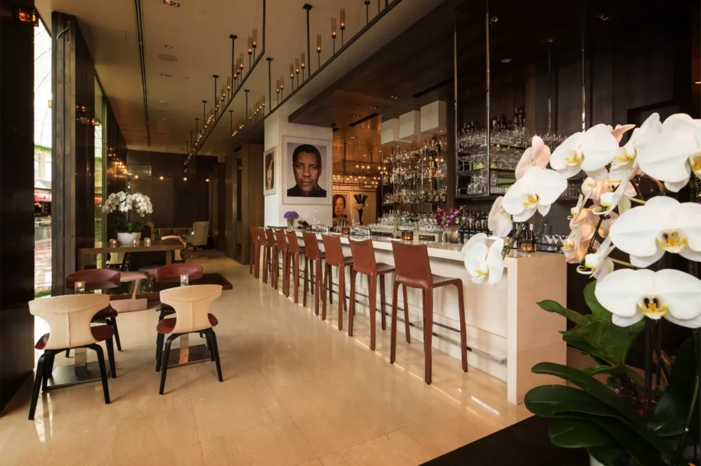 An eye-level shot captures a modern, luxury bar lounge featuring a long marble counter lined with high-backed brown leather barstools. The sophisticated interior is illuminated by unique pendant lighting and decorated with large white orchid arrangements in the foreground, with portrait artwork on the wall behind the bar.
