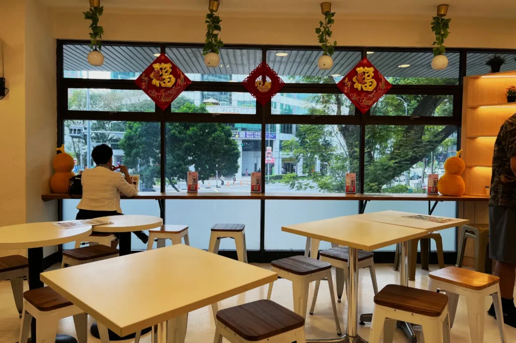 A cozy cafe with white tables and wooden stools facing large windows. Red decorations hang above, and a person sits at the counter, creating a serene atmosphere.