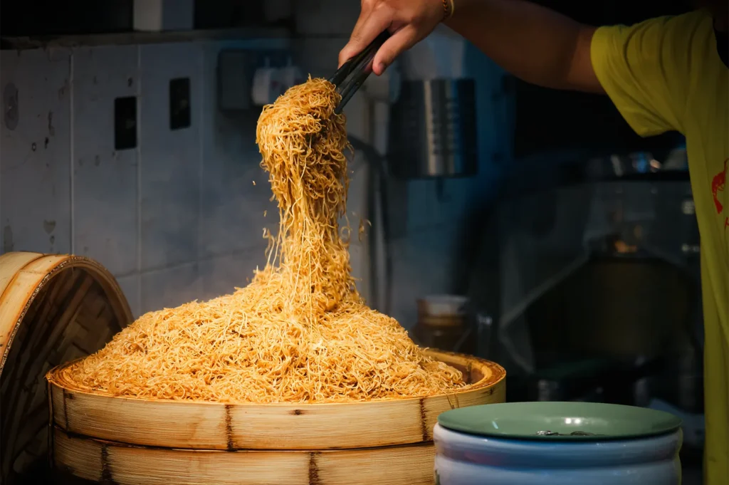 A close-up shot captures a vendor using metal tongs to lift a steaming portion of thin, seasoned noodles from a large, heaped pile inside a round bamboo steamer basket. The background shows a simple, tiled kitchen stall setting with a green serving plate ready in the foreground.