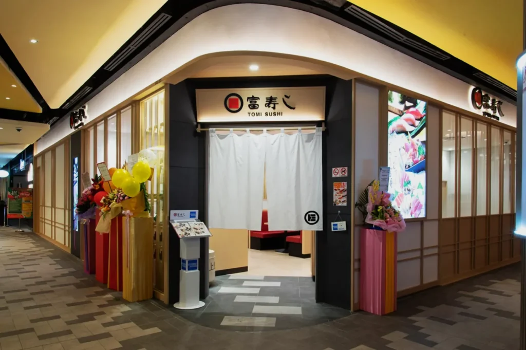 Front entrance of Tomi Sushi restaurant with a white noren curtain, surrounded by colorful flower arrangements, in a modern indoor setting.