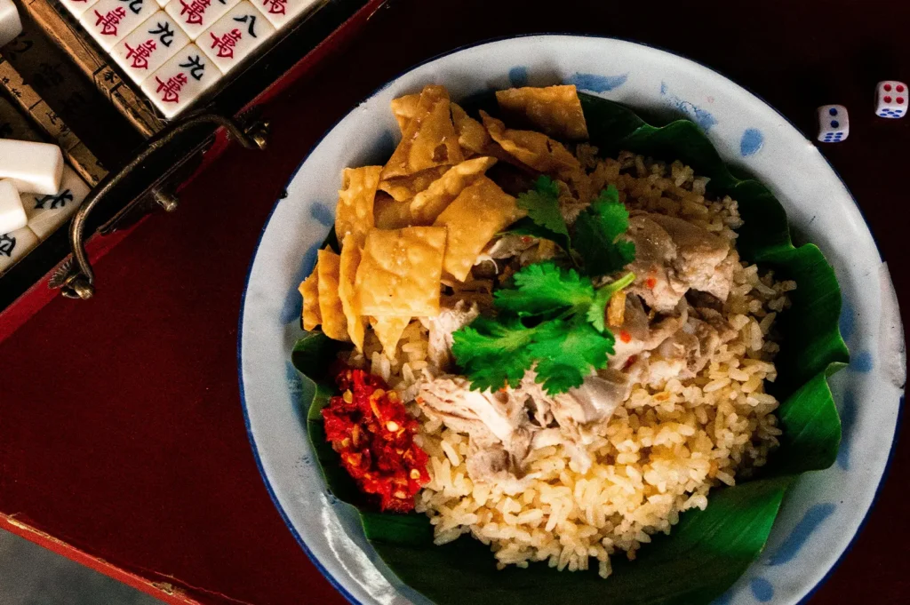 A top-down shot captures a bowl of Hainanese chicken rice served on a vibrant green banana leaf, topped with tender sliced chicken, fresh cilantro, crispy wonton skins, and a side of bright red chili paste. The bowl sits on a red tabletop next to scattered mahjong tiles and a pair of dice, creating a culturally rich setting.