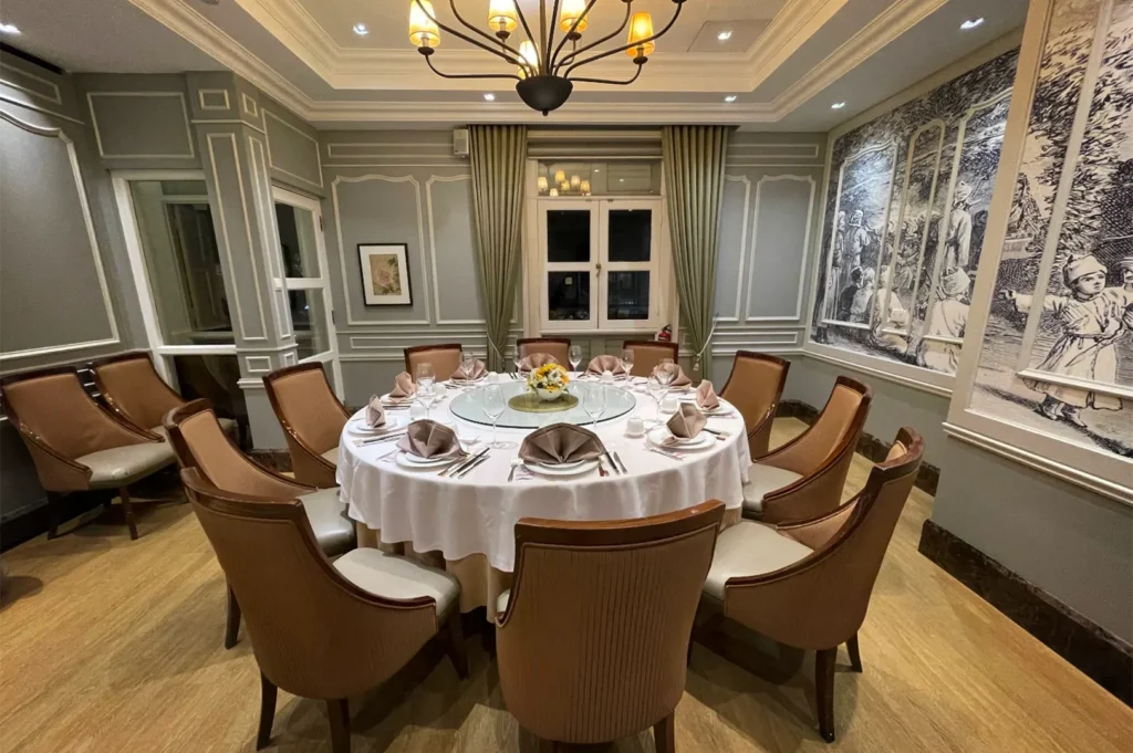 An eye-level shot captures a private dining room featuring a large, round table meticulously set with a white tablecloth, formal dinnerware, folded brown napkins, and a central Lazy Susan. The sophisticated space is adorned with light blue paneled walls, a large black-and-white mural of traditional figures, light wood flooring, and an elegant multi-bulb chandelier hanging overhead.