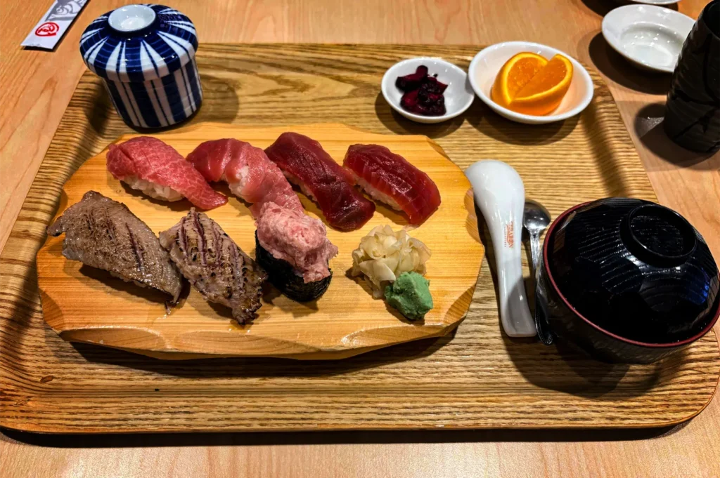 A high-angle shot captures a wooden platter of sushi served on a matching wooden tray alongside side dishes and soup. The platter holds an assortment of tuna nigiri ranging from light pink to deep red, two pieces of seared aburi sushi, a gunkan maki roll, pickled ginger, and a dollop of green wasabi.