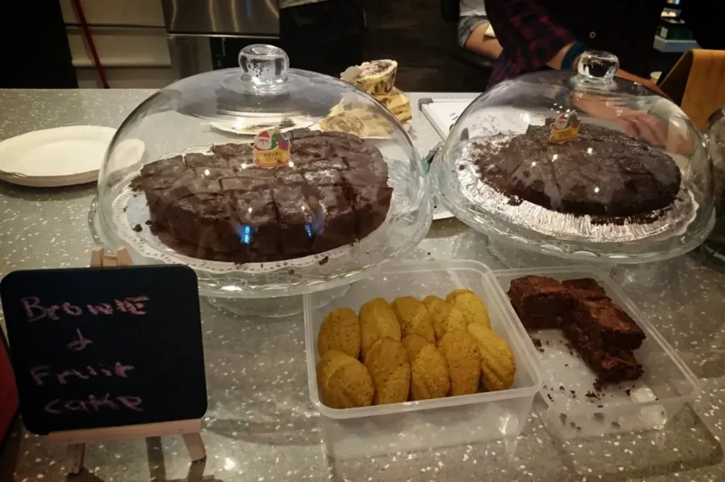 An eye-level, close-up shot captures a selection of baked goods arranged on a speckled grey countertop. In the center, two large chocolate cakes or brownie slabs rest on glass cake stands under clear plastic domes, each topped with a small, festive holiday toothpick. In front of them sit two clear plastic containers: the left holds neatly lined oval cookies or madeleines, and the right contains several square brownie pieces. To the far left, a small black chalkboard sign mounted on a wooden easel displays the handwritten text "Brownie & Fruit Cake" in pink chalk, while a large glass jar labeled "Free cookies" is partially visible on the right next to a person wearing a red and black plaid shirt.