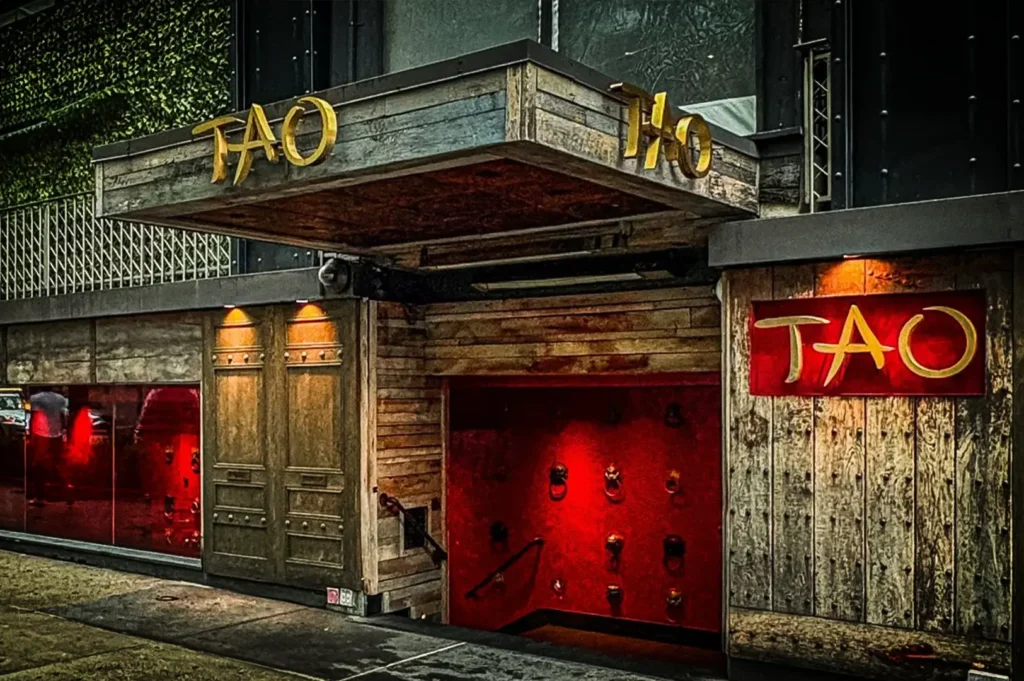A medium, low-angle shot captures the distinctive exterior entrance of TAO restaurant along a city sidewalk. The facade is constructed from rustic, weathered wood panels and heavy metal detailing, featuring a large wooden overhang with the word "TAO" mounted in bold gold lettering on both the front and side. Below the canopy, a wide stairwell painted a vibrant, glowing red leads downward, its walls studded with rows of ornamental metal door knockers. To the right of the stairs, a red rectangular sign displays the stylized gold "TAO" logo, while a glass window to the far left reveals a glimpse of a brightly lit red interior, altogether creating a dramatic and welcoming urban entrance.