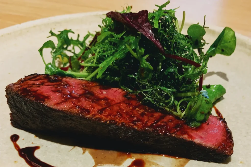 A seared steak, cooked medium-rare, topped with a mound of fresh mixed greens drizzled in a glossy sauce on a beige plate, creating a gourmet presentation.
