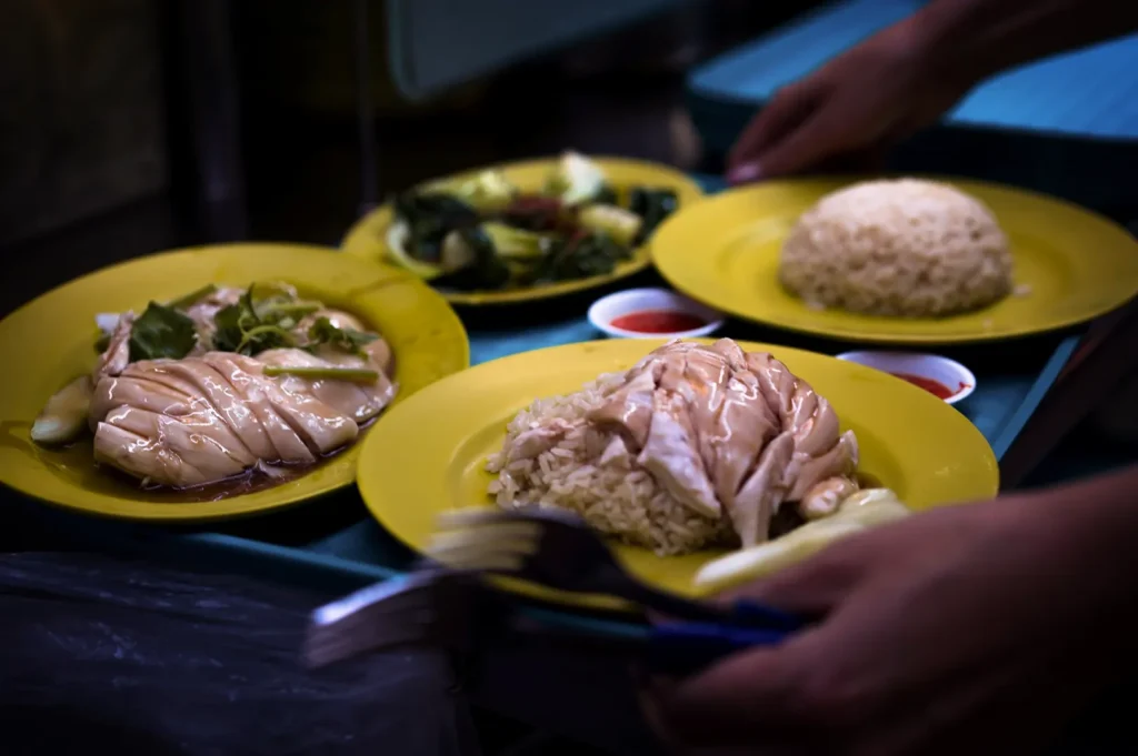 A close-up shot captures a tray of Hainanese chicken rice served on vibrant yellow plates, featuring sliced poached chicken over seasoned rice alongside steamed greens and small saucers of chili sauce. A person's hands are seen holding the blue tray, highlighted by a shallow depth of field.