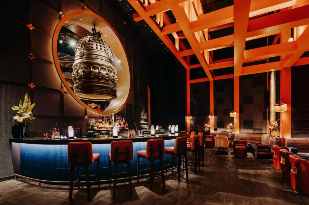 Stylish bar featuring a blue counter and red stools, with a large ornamental sculpture in a circular frame. Warm lighting creates an inviting, modern vibe.