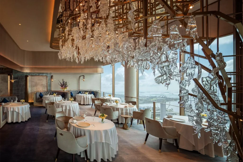 An eye-level shot captures a luxurious, high-rise restaurant dining room featuring round tables with white tablecloths and plush armchairs arranged near floor-to-ceiling windows. A massive, intricate glass sculpture chandelier hangs from the ceiling, reflecting light above the space while offering guests a panoramic view of a coastal city and waterfront below.