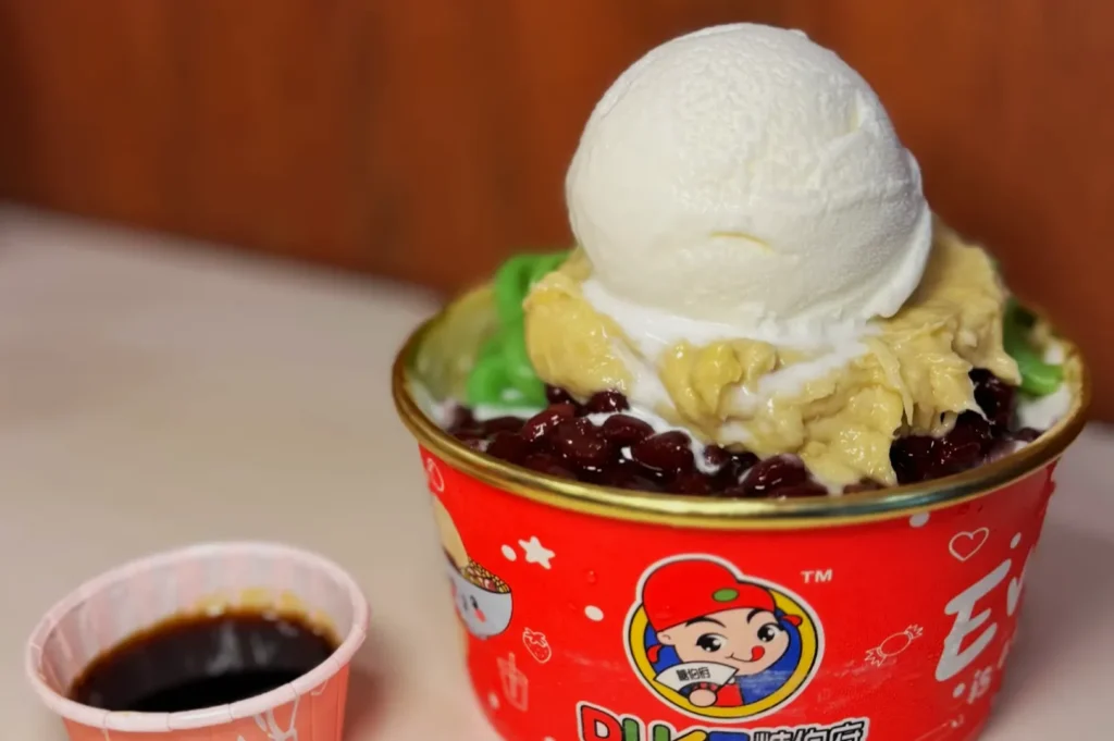 A bowl of shaved ice dessert with a scoop of vanilla ice cream, red beans, and green syrup, in a red cup with cartoon graphics, next to a small cup of dark syrup.