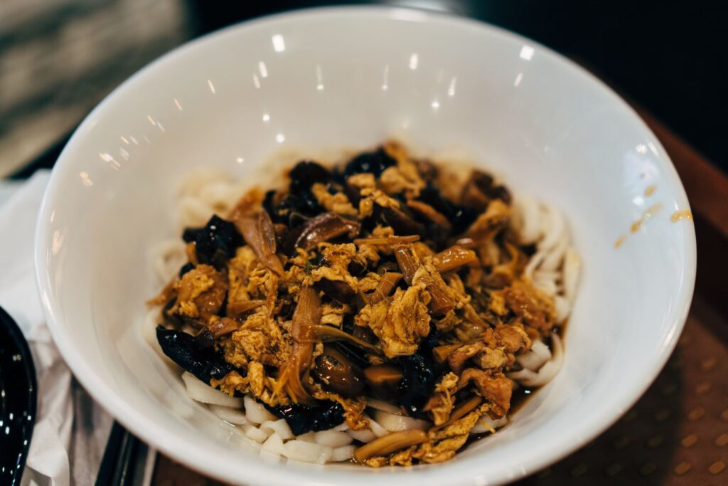 A close-up shot captures a deep white ceramic bowl filled with thick, flat noodles topped with a savory mixture of scrambled egg, wood ear mushrooms, and shredded meat in a dark sauce. The dish sits on a dark tabletop with chopsticks partially visible in the blurred foreground.