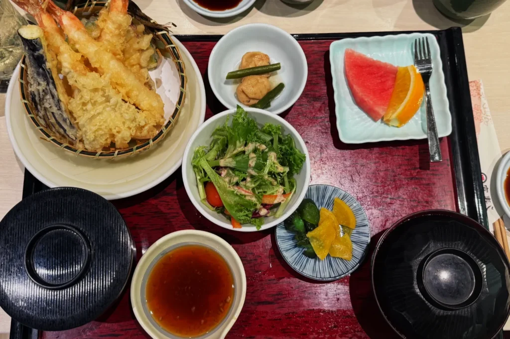A Japanese meal on a red tray includes tempura shrimp, mixed salad, pickles, tofu, dipping sauce, miso soup, and fresh fruit slices.