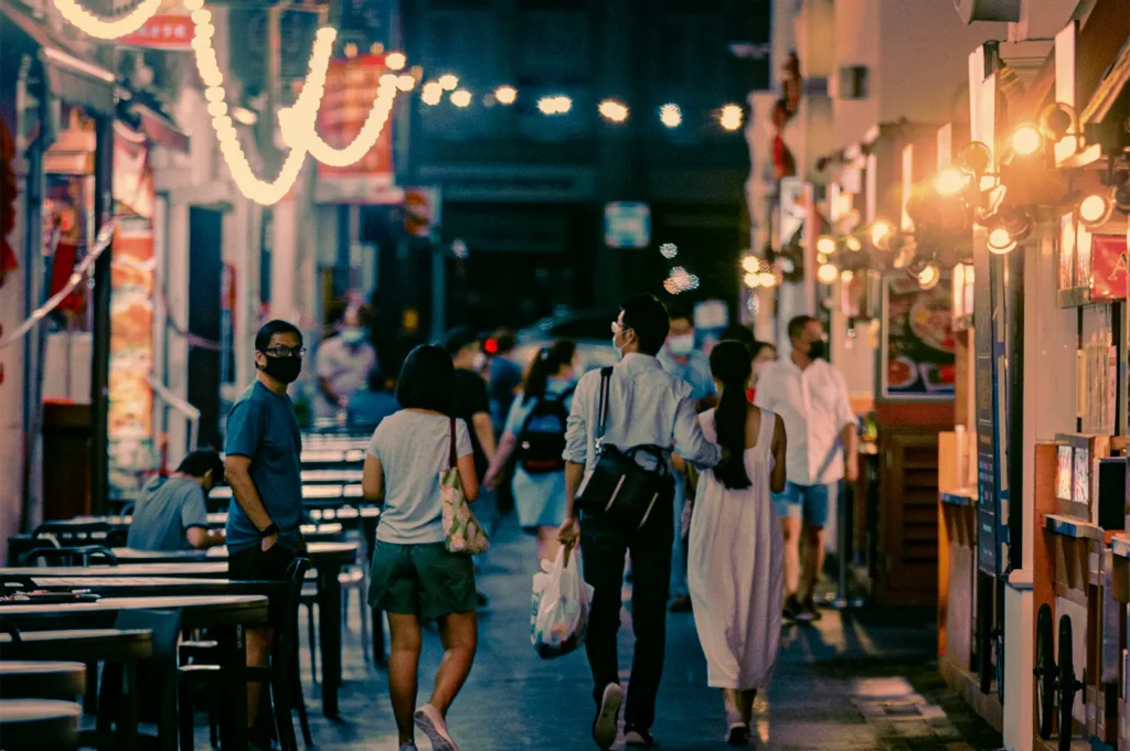An eye-level shot captures pedestrians walking down a lively city street at night, with a couple seen from behind carrying a shopping bag as they stroll past outdoor restaurant seating. Warm string lights and illuminated storefronts line the narrow lane, casting a cozy glow over the passersby, several of whom are wearing face masks.