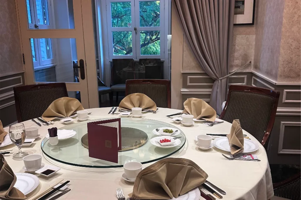 An eye-level shot captures a large, round dining table inside a private restaurant room, meticulously set with a white tablecloth, formal dinnerware, and beige fan-folded napkins around a central glass Lazy Susan holding small appetizers and a red menu. The elegant space features classic wainscoted walls, high-backed wooden chairs, and a half-open glass door in the background revealing a glimpse of green trees outside.