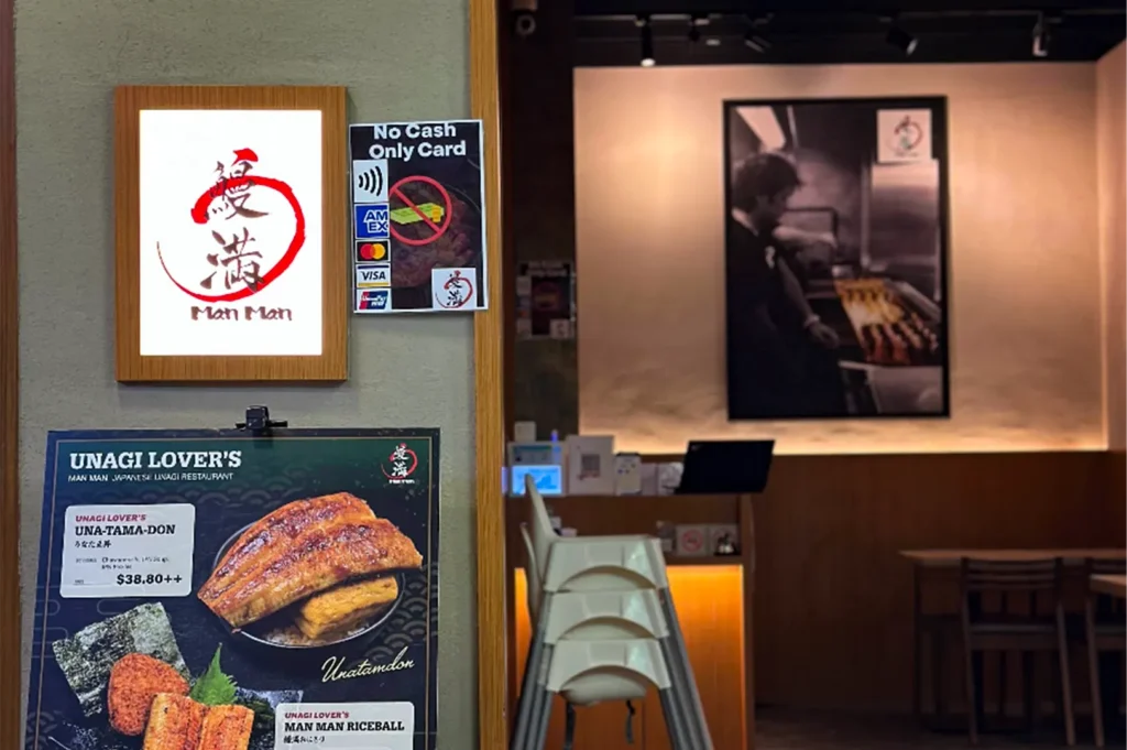 An eye-level shot shows the entrance of Man Man Japanese Unagi Restaurant, featuring a lit logo sign, a card-only payment notice, and a menu board advertising their Una-Tama-Don. Through the open doorway, the restaurant interior is visible with stacked high chairs, a counter, and a large black-and-white poster of a chef grilling eel on the back wall.