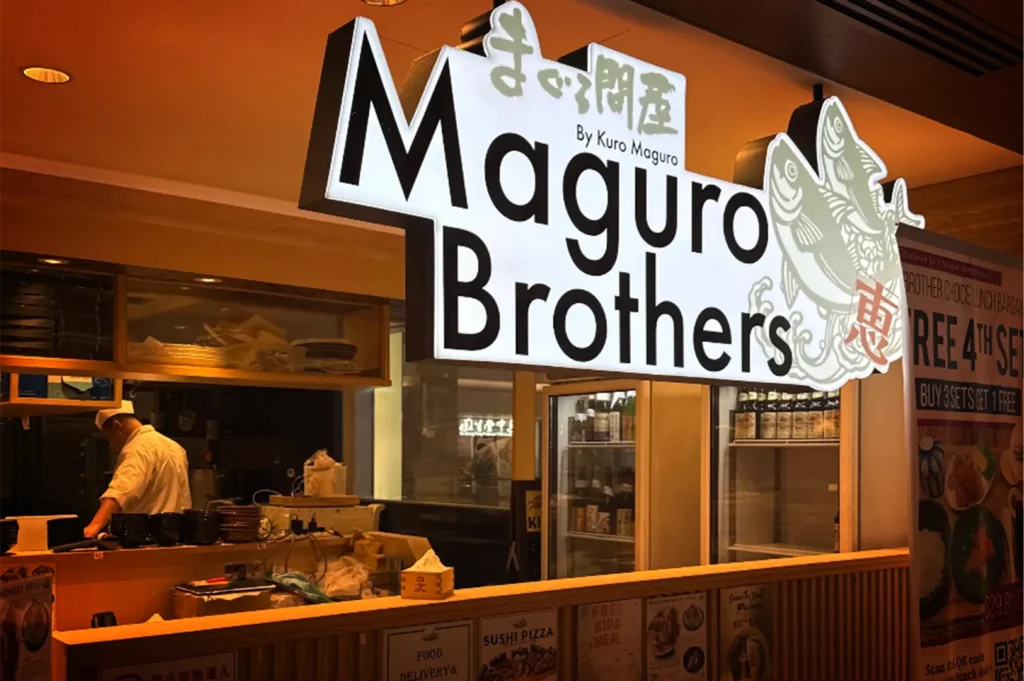 An eye-level shot captures the storefront of a restaurant named "Maguro Brothers by Kuro Maguro," highlighted by a large, illuminated white sign with bold black text and a fish illustration. Below the signage, a chef in a white uniform can be seen working in the dimly lit, open kitchen area behind a wooden counter.
