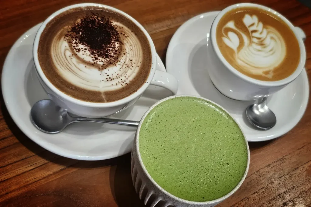 A high-angle, close-up shot captures a trio of beautifully crafted café beverages clustered together on a warm, grained wooden table. On the upper left, a large white ceramic cup of cappuccino or hot chocolate features a rich dusting of cocoa powder over swirling white foam, resting on a matching white saucer with a small silver spoon. To the upper right, a smaller white cup displays a latte or flat white decorated with elegant swan latte art in the creamy microfoam, also paired with its own saucer and spoon. In the foreground, a textured, handleless beige ceramic cup is filled to the brim with a vibrant, finely frothed green matcha latte, completing the cozy drink arrangement.
