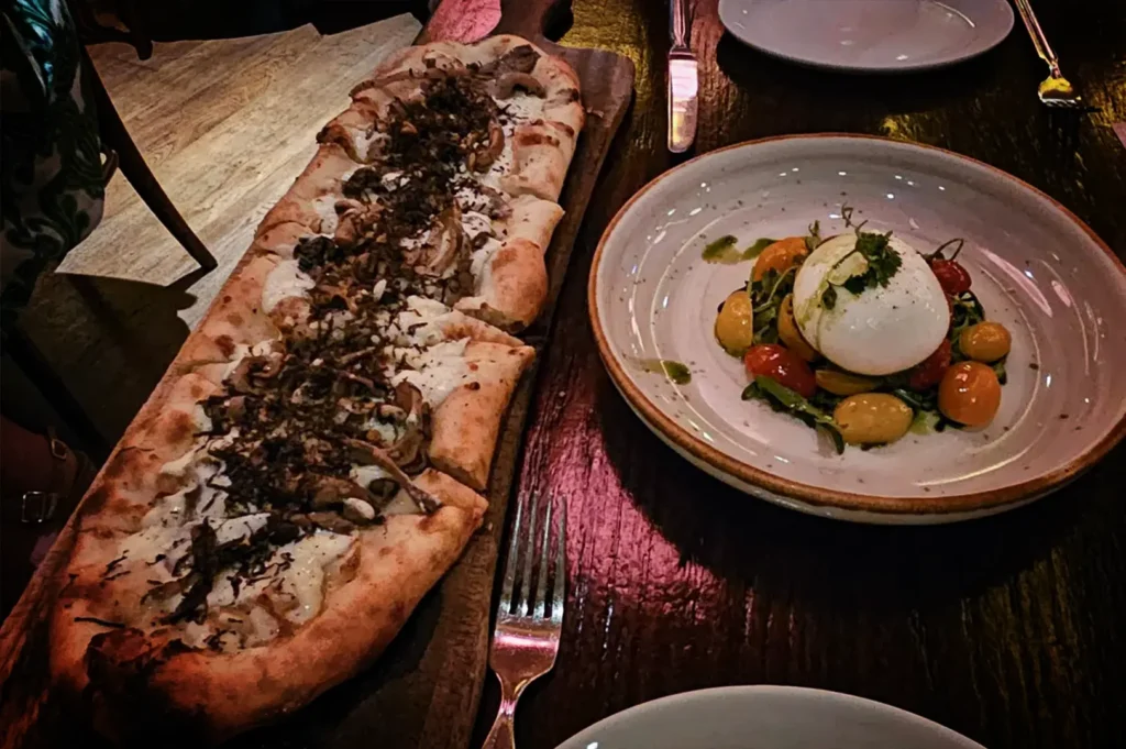 A high-angle, close-up shot captures an appetizing meal served on a dark wooden table in a restaurant with warm, moody lighting. On the left, a long, rustic oval flatbread pizza heavily topped with melted white cheese, mushrooms, and herbs is presented on a curved wooden serving board. To the right, a round ceramic plate holds a large ball of white burrata cheese drizzled with pesto, resting on a vibrant bed of red and yellow cherry tomatoes and fresh arugula. Silverware, drinking glasses, and a small, glowing votive candle holder are scattered around the dishes, completing the intimate dining setting.