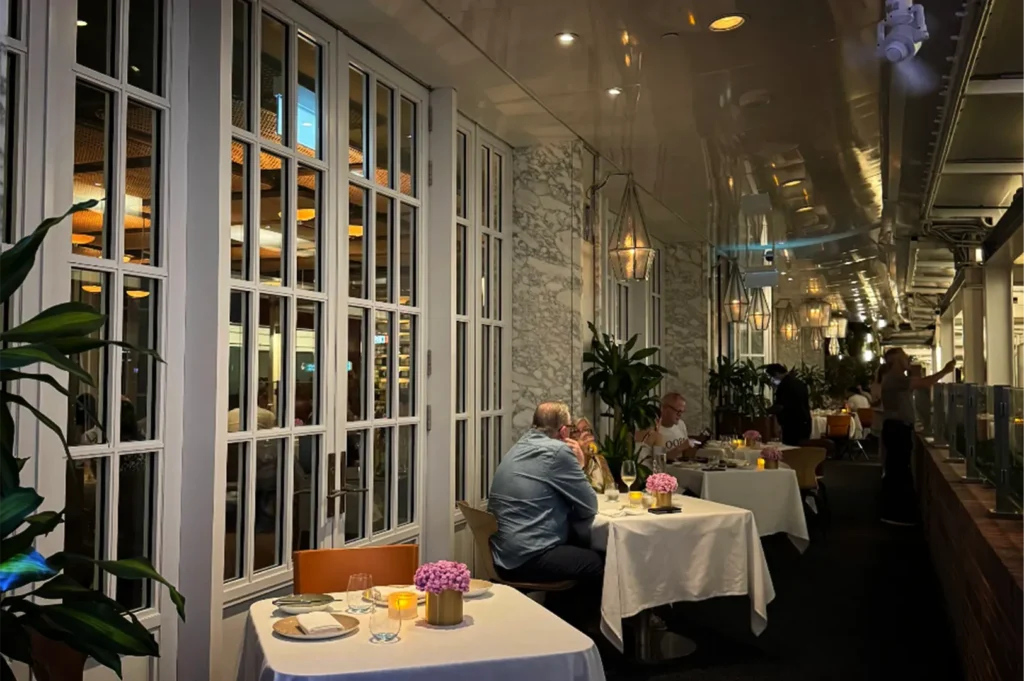 A medium, eye-level shot captures a lively outdoor balcony or covered terrace dining area of an upscale restaurant at night. On the left, a long wall features large, white multi-paned French doors and windows that reveal a warmly lit indoor dining room, accented by lush green potted plants in the foreground. Several tables dressed in crisp white tablecloths run down the length of the terrace, occupied by diners engaged in conversation, while a line of geometric, warm-toned lantern light fixtures hangs from the sleek, reflective ceiling overhead. In the background, restaurant staff move between tables, and a low glass-and-wood partition borders the right edge of the narrow dining walkway.