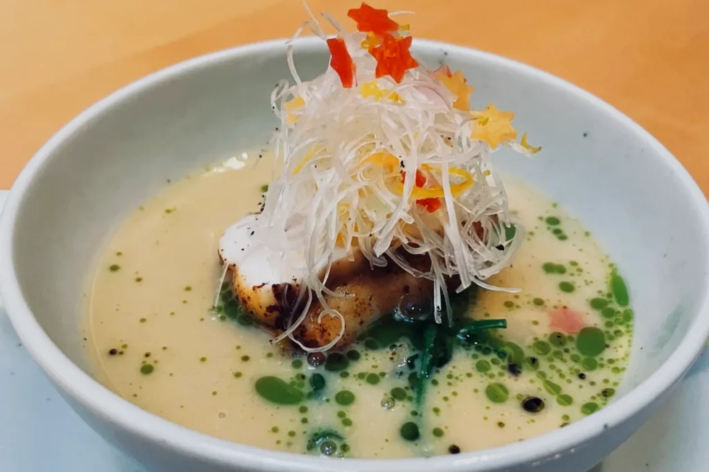 A bowl of creamy soup with grilled fish, topped with shredded radish and colorful flower petals. Green herbs add a fresh touch. Elegant and appetizing.