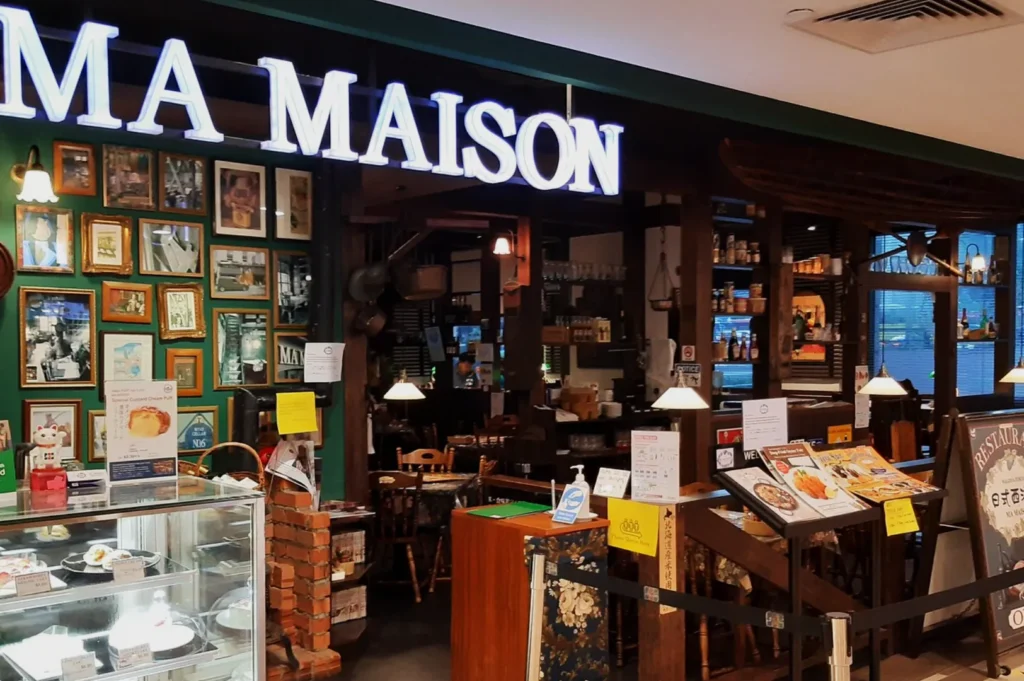 Restaurant entrance with the illuminated sign "MA MAISON" in a cozy, rustic setting. Features framed art on a green wall, warm lighting, and a pastry display.