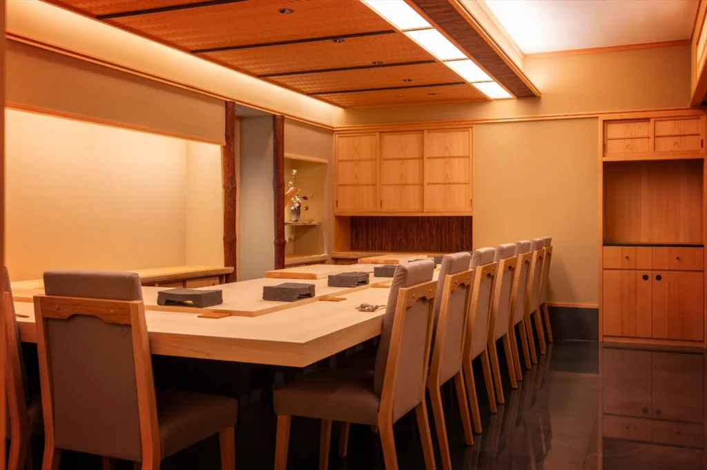 An eye-level shot captures a minimalist Japanese-style dining room featuring a long, light wood counter lined with high-backed, upholstered dining chairs. The warm, serene space is illuminated by soft, recessed lighting and decorated with traditional wooden cabinetry and paneling.