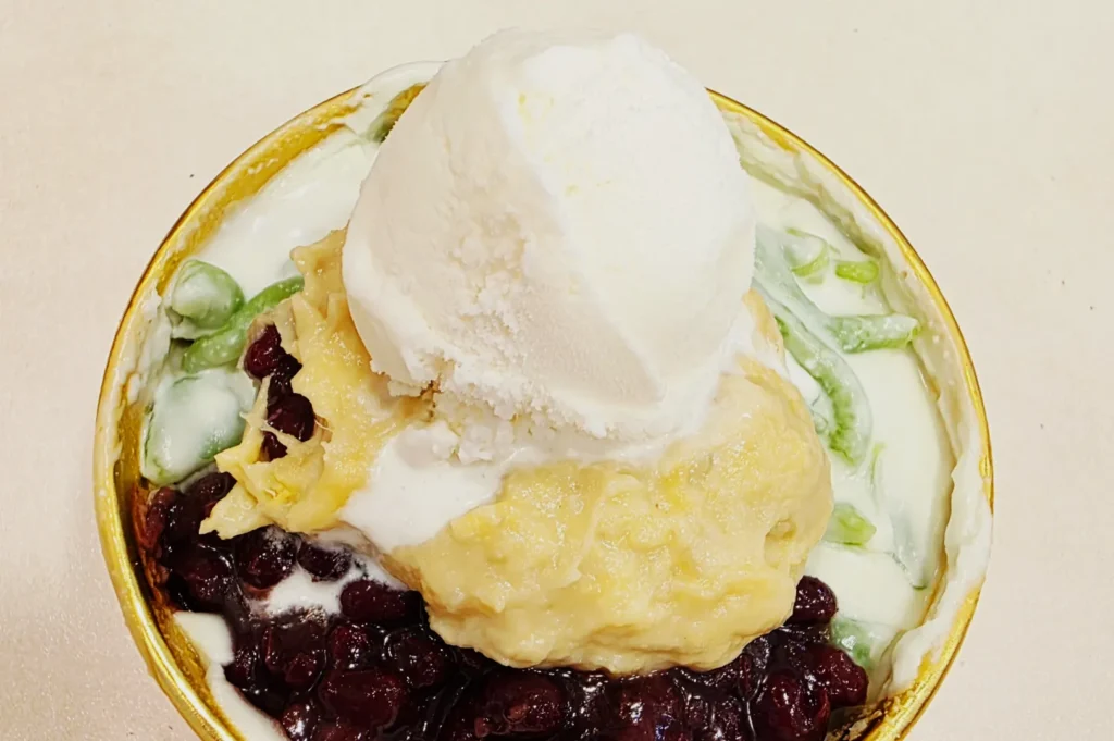 A vibrant bowl of halo-halo dessert with creamy shaved ice, green jelly, red beans, banana, and topped with a generous scoop of vanilla ice cream.