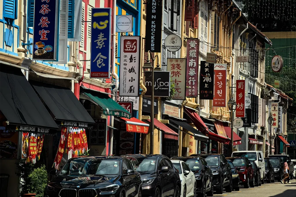 A vibrant, eye-level shot captures a row of colorful, traditional shophouses lining a city street filled with parked cars. The buildings feature colorful facades with hanging awnings and a dense cluster of vertical business signs written in both Chinese characters and English.