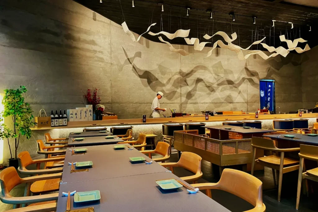 Modern Japanese restaurant interior with wooden chairs, long tables set with chopsticks and square plates, a chef working, and sculptural ceiling decor.