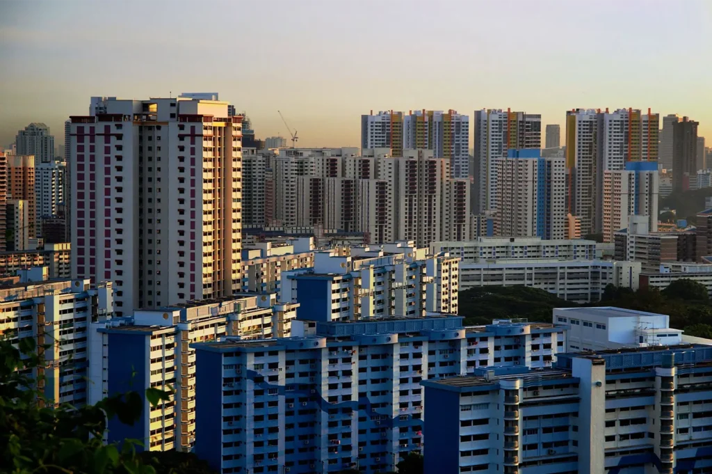 A high-angle cityscape view showcases a dense grid of public housing apartment blocks painted in various shades of blue, white, and orange accents. The warm, golden sunlight of early morning or late afternoon casts long shadows across the buildings, highlighting the tightly packed urban architecture against a pale sky.