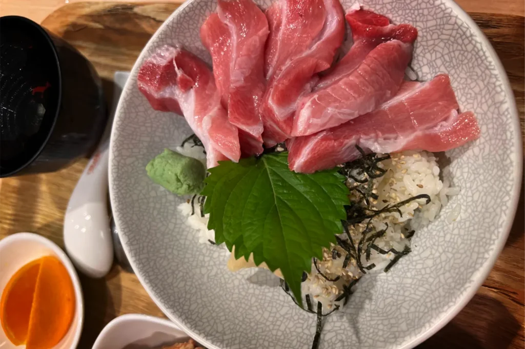 A top-down shot captures a maguro donburi bowl filled with thick, marbled pink slices of fresh tuna arranged over a bed of seasoned rice. The dish is garnished with a vibrant green shiso leaf, shredded nori, sesame seeds, and a dollop of wasabi, served on a wooden tray with small side dishes.