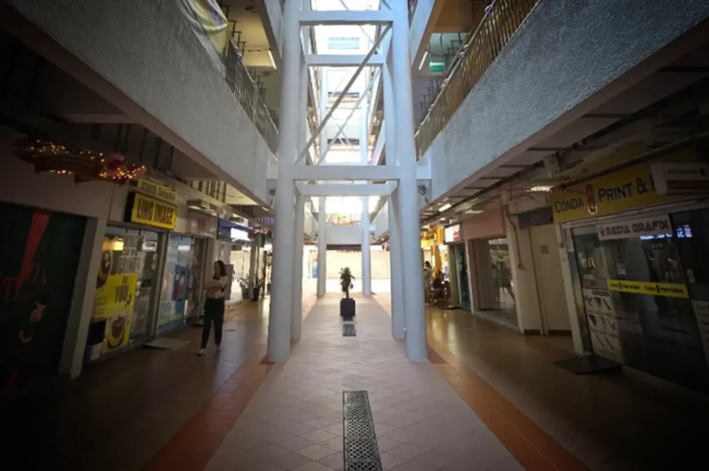 A wide, eye-level shot captures the interior courtyard and walkways of a multi-story commercial complex or housing block, featuring a prominent white structural framework of pillars and beams running down the center. On the ground level, tiled walkways line both sides of the central atrium, passing by storefronts with signs for businesses such as "LING IMAGE" on the left and "CONDA PRINT & MEDIA GRAFX" on the right. A few scattered pedestrians stand or walk near the shops under the shadow of the upper floors, while natural light streams down from the open air space above the central structural pillars.