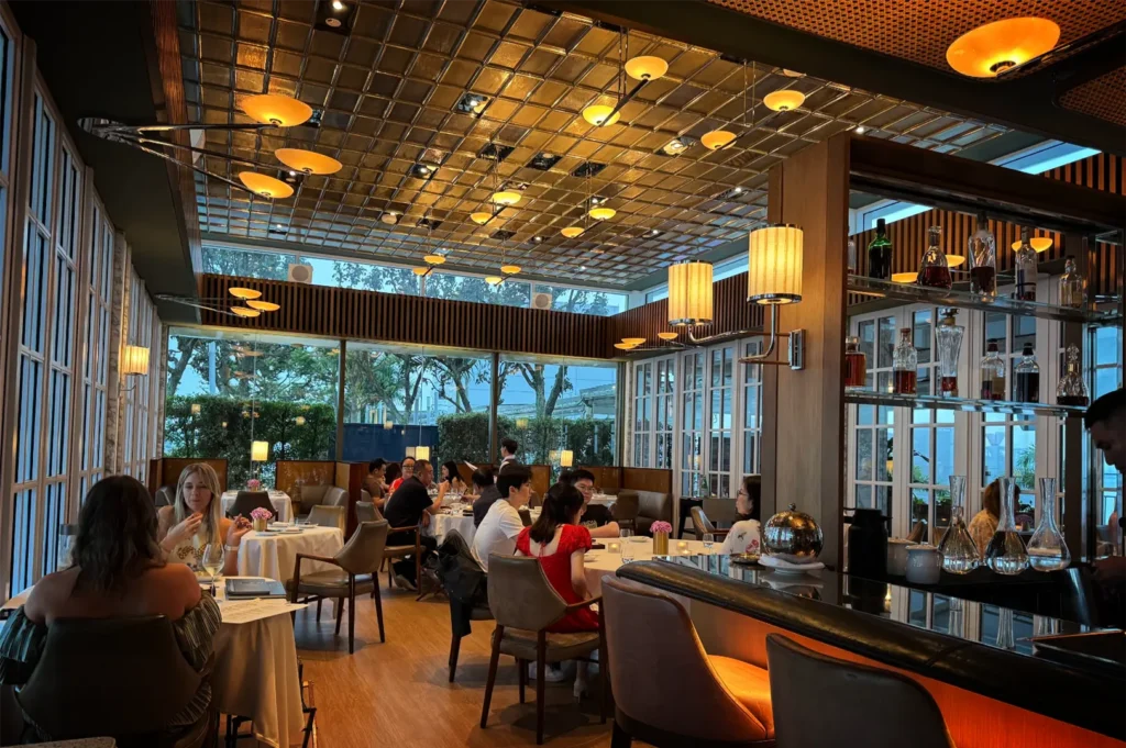 A wide, eye-level shot captures the warm and bustling atmosphere of a high-end restaurant dining room filled with patrons. On the right, a sleek black bar counter curves into the foreground, illuminated by orange under-lighting, with a glass partition display case behind it showcasing liquor bottles and glass decanters. Multiple groups of diners are seated at tables draped in crisp white tablecloths across the smooth wooden floor, engaged in conversation. The room features a striking grid-patterned metallic ceiling accented by numerous warm, disc-shaped pendant lights, while large floor-to-ceiling glass windows at the back look out onto an illuminated terrace with green trees during dusk.